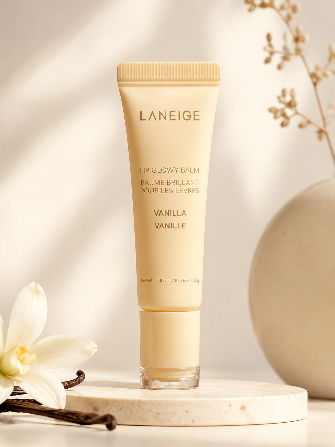 A tube of LANEIGE Lip Glowy Balm in Vanilla is displayed against a soft background, accompanied by a white flower and a round stone, highlighting its elegant packaging.