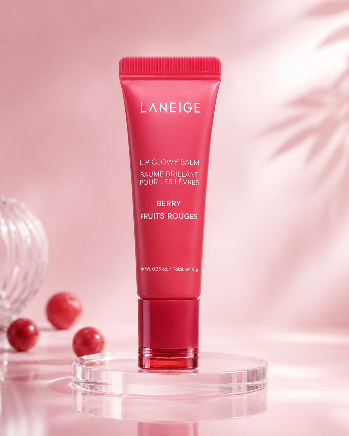 A tube of Laneige Lip Glowy Balm in Berry sits on a clear circular pedestal. The background features soft pink tones, enhancing the products vibrant packaging.