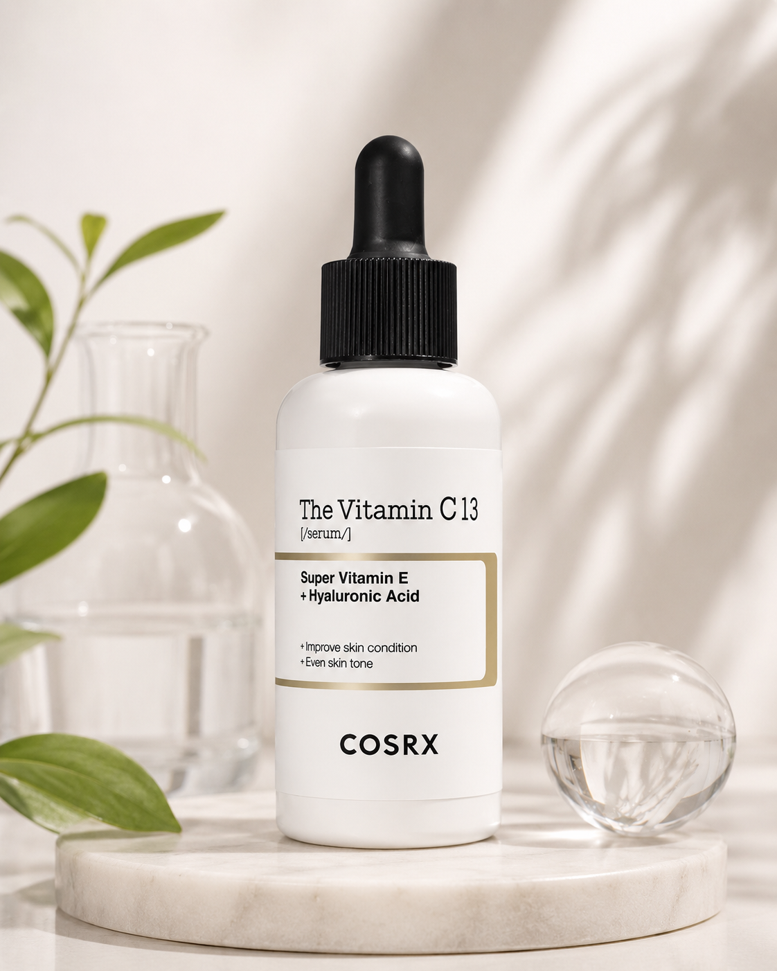 COSRX The Vitamin C 13 Serum displayed on a light stone pedestal with fresh greenery, glass accents and soft natural light, creating a clean, bright skincare aesthetic that reflects its radiance-focused formula.