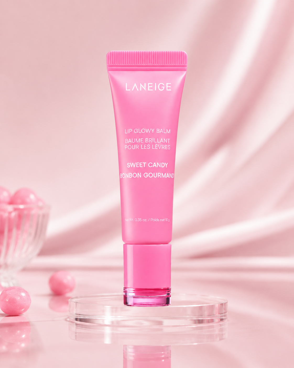 A pink tube of LANEIGE Lip Glowy Balm in Sweet Candy sits atop a clear, reflective surface, surrounded by glossy pink candy-like spheres. The backdrop features a soft, pastel aesthetic.